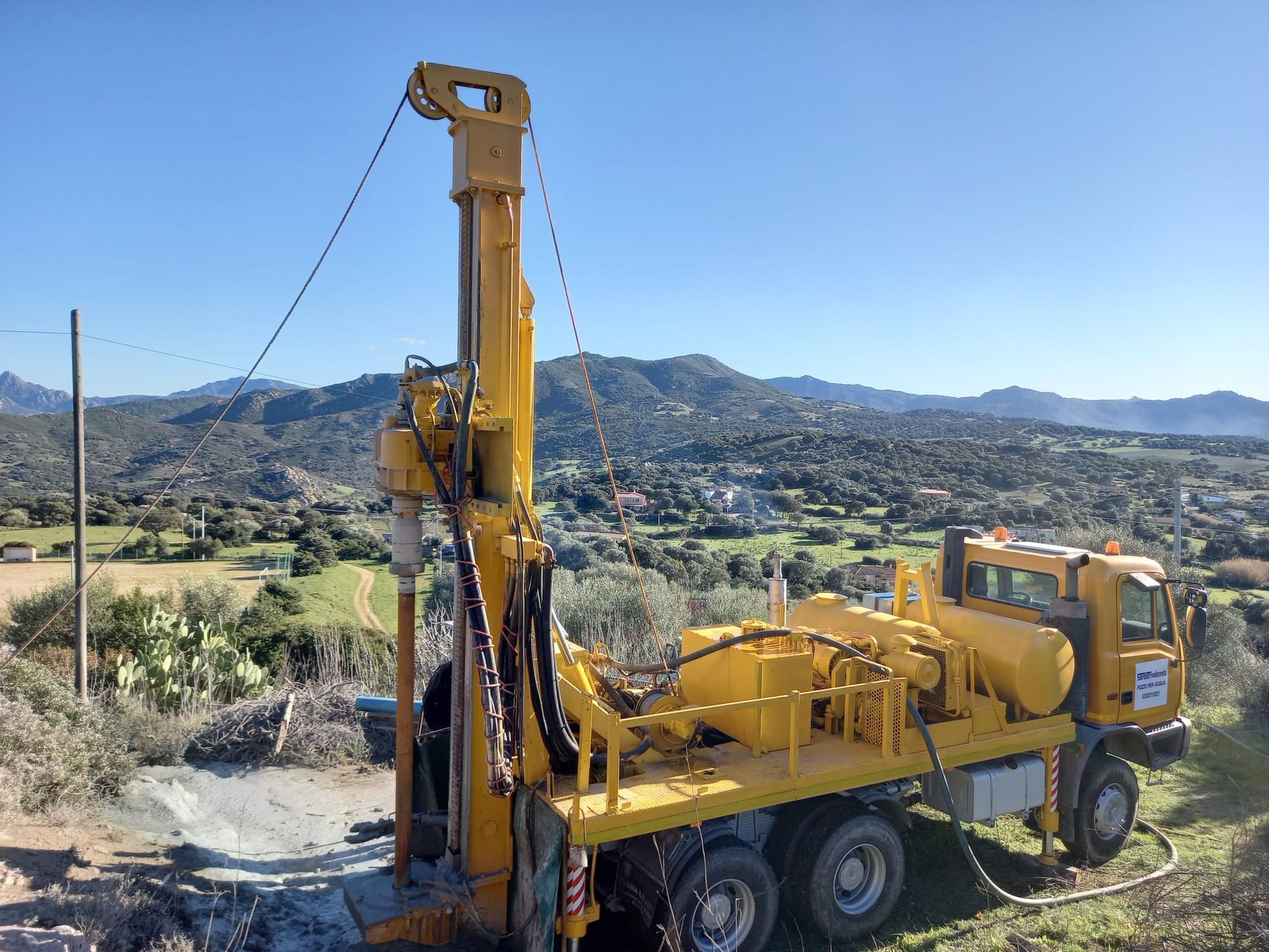 Una piattaforma di perforazione gialla su un camion in un paesaggio rurale, mentre perfora un foro sotto un cielo azzurro e terso Sarda Trivelsonda Group Olbia  Loiri Porto San Paolo - Loiri (SS)