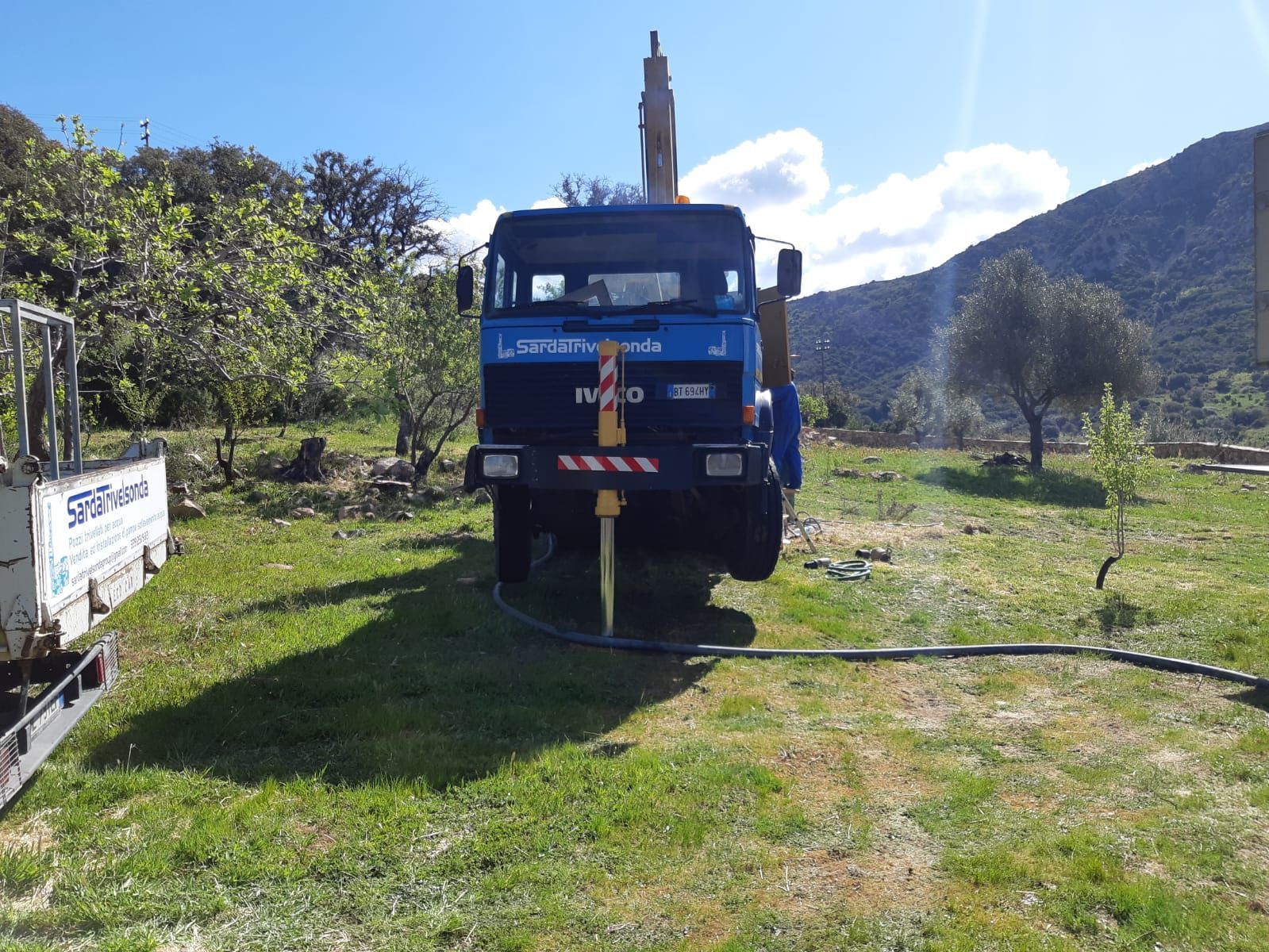 Camion perforatore blu sull'erba in un campo, con una montagna sullo sfondo Sarda Trivelsonda Group Olbia  Loiri Porto San Paolo - Loiri (SS)