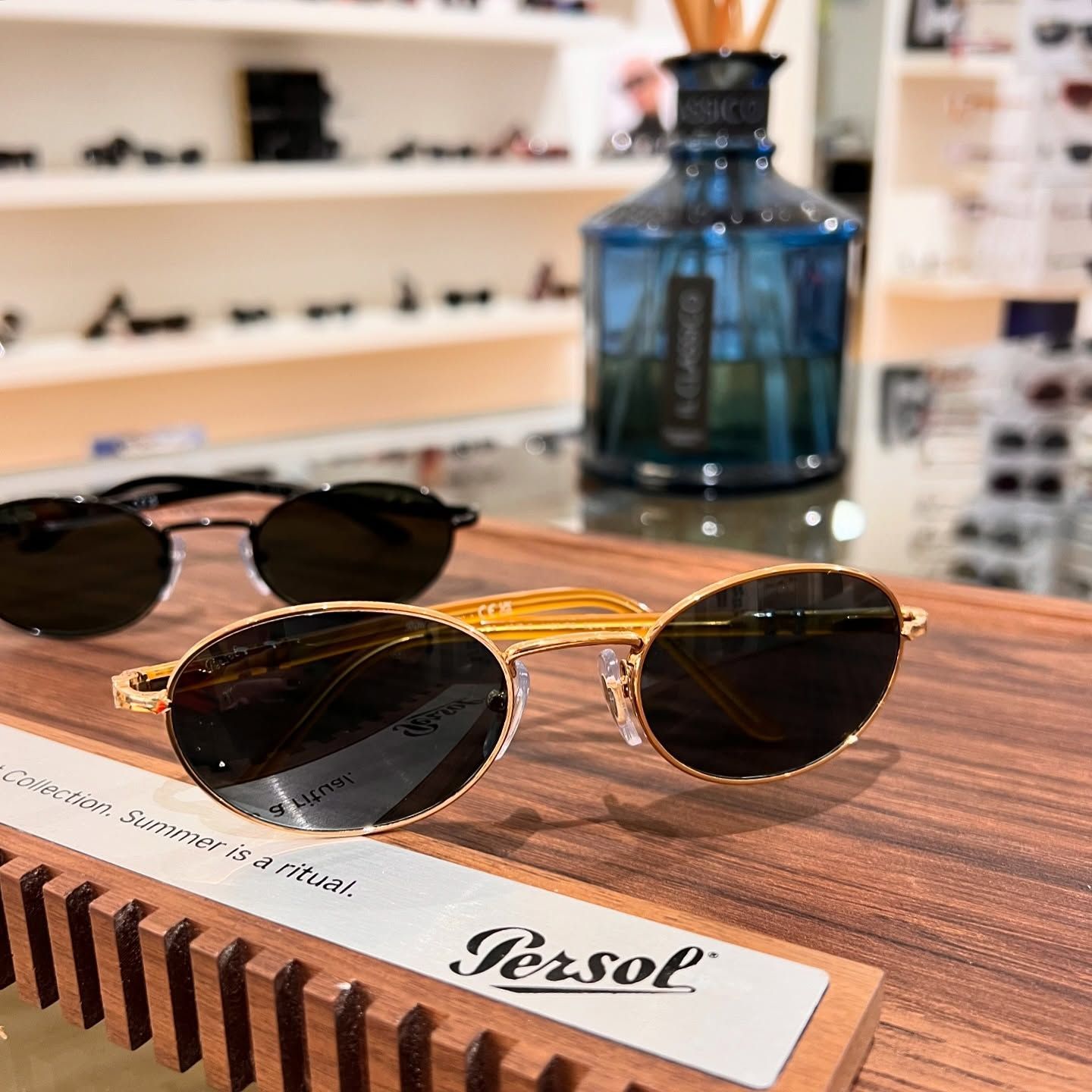 Occhiali da sole Persol con montatura dorata su un espositore in legno in un negozio di ottica, con un diffusore a canne blu sullo sfondo.