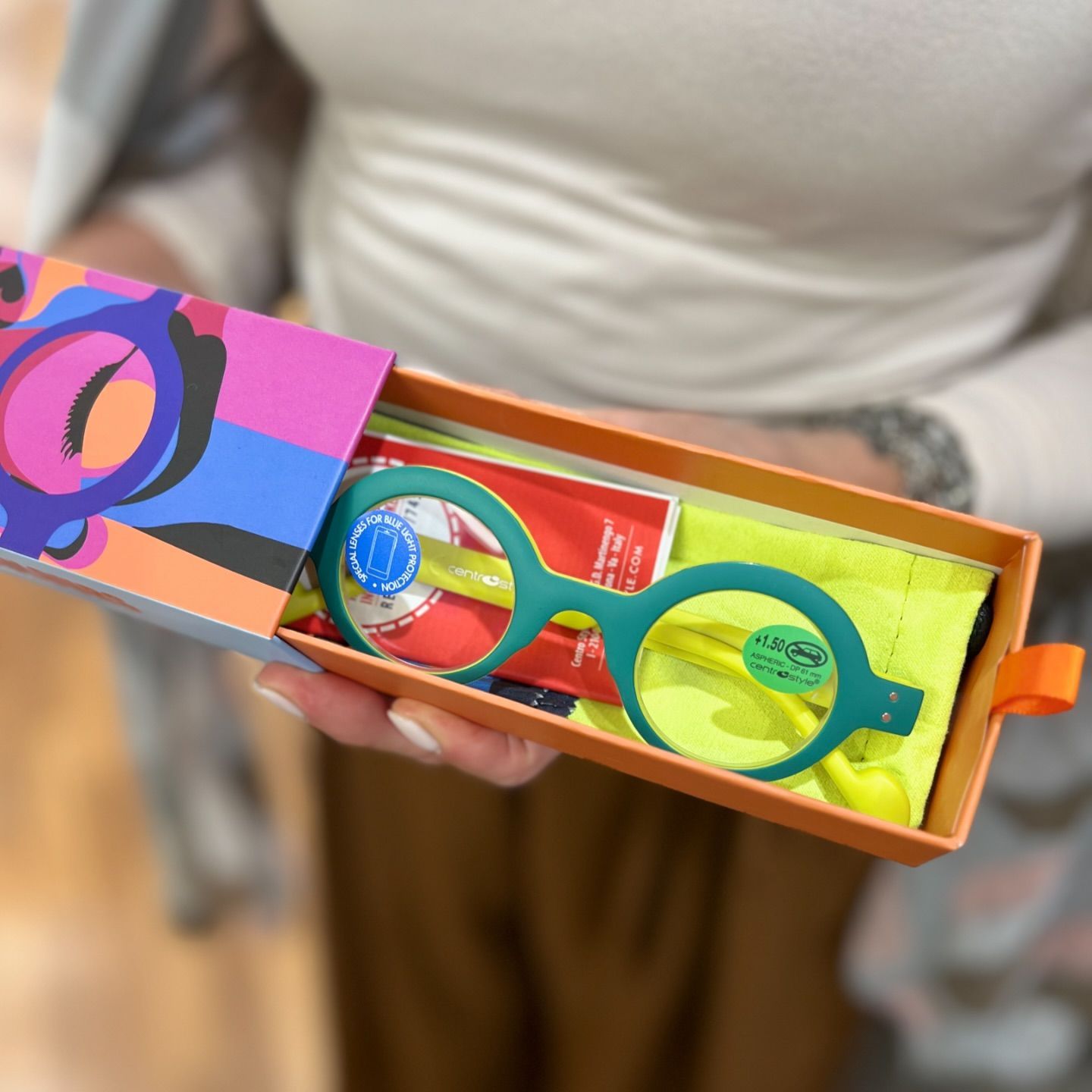 Una persona tiene in mano una scatola scorrevole aperta e colorata, contenente bicchieri rotondi color verde acqua brillante con dettagli gialli.