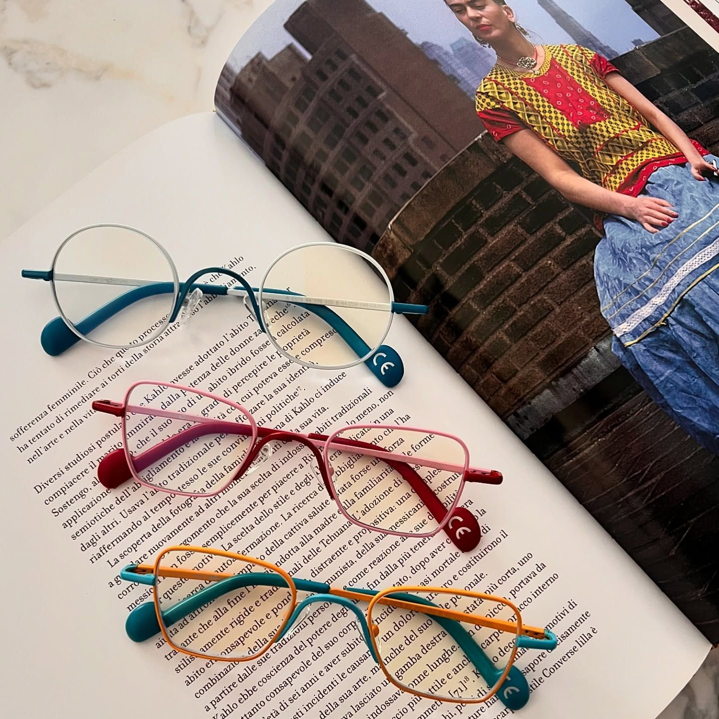 Tre paia di occhiali colorati con montature rotonde, a occhi di gatto e ovali poggiano su un libro aperto che contiene un ritratto.