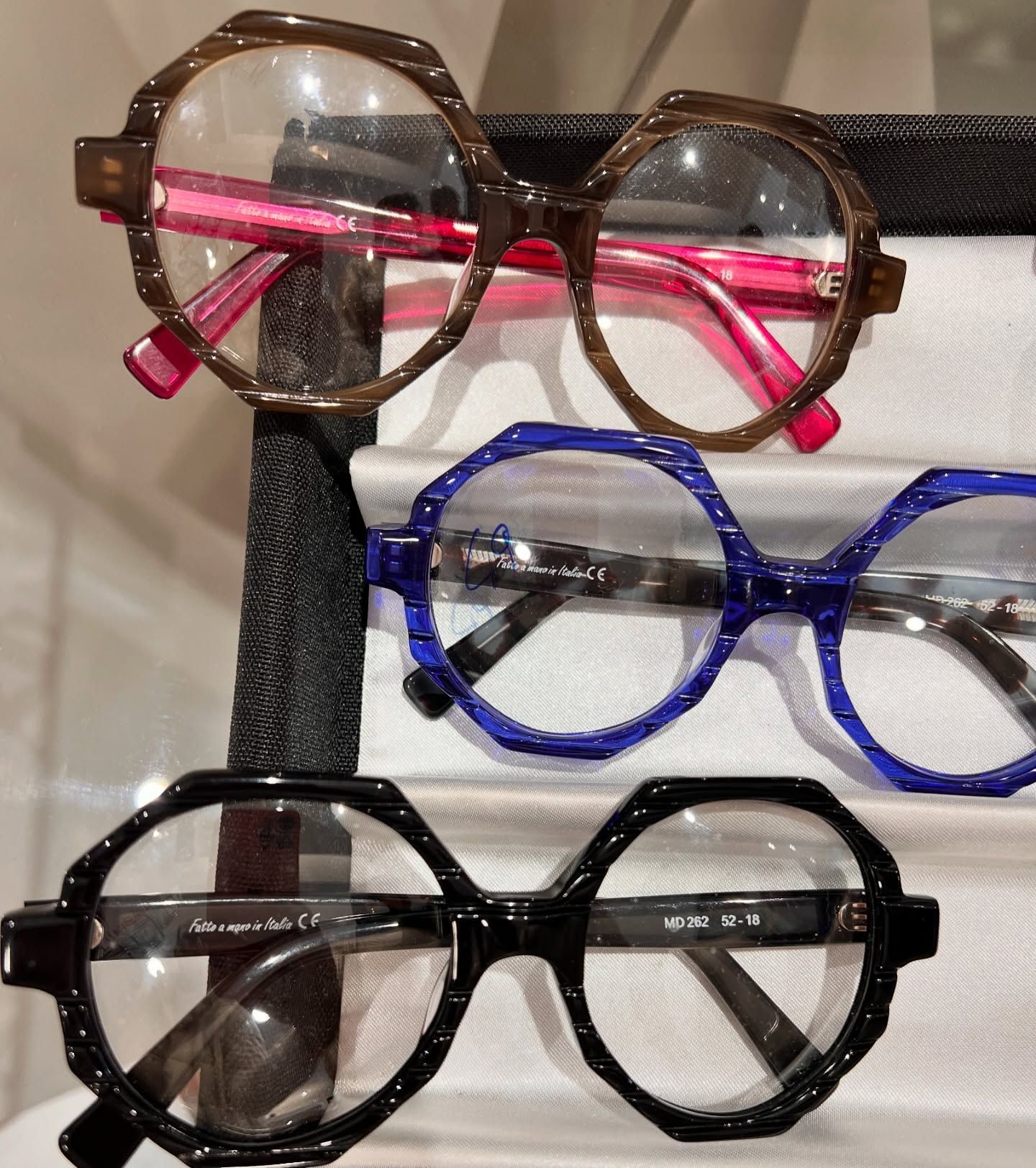 Tre paia di occhiali con montatura ottagonale impilati verticalmente, di colore marrone con aste rosa, blu e nero.