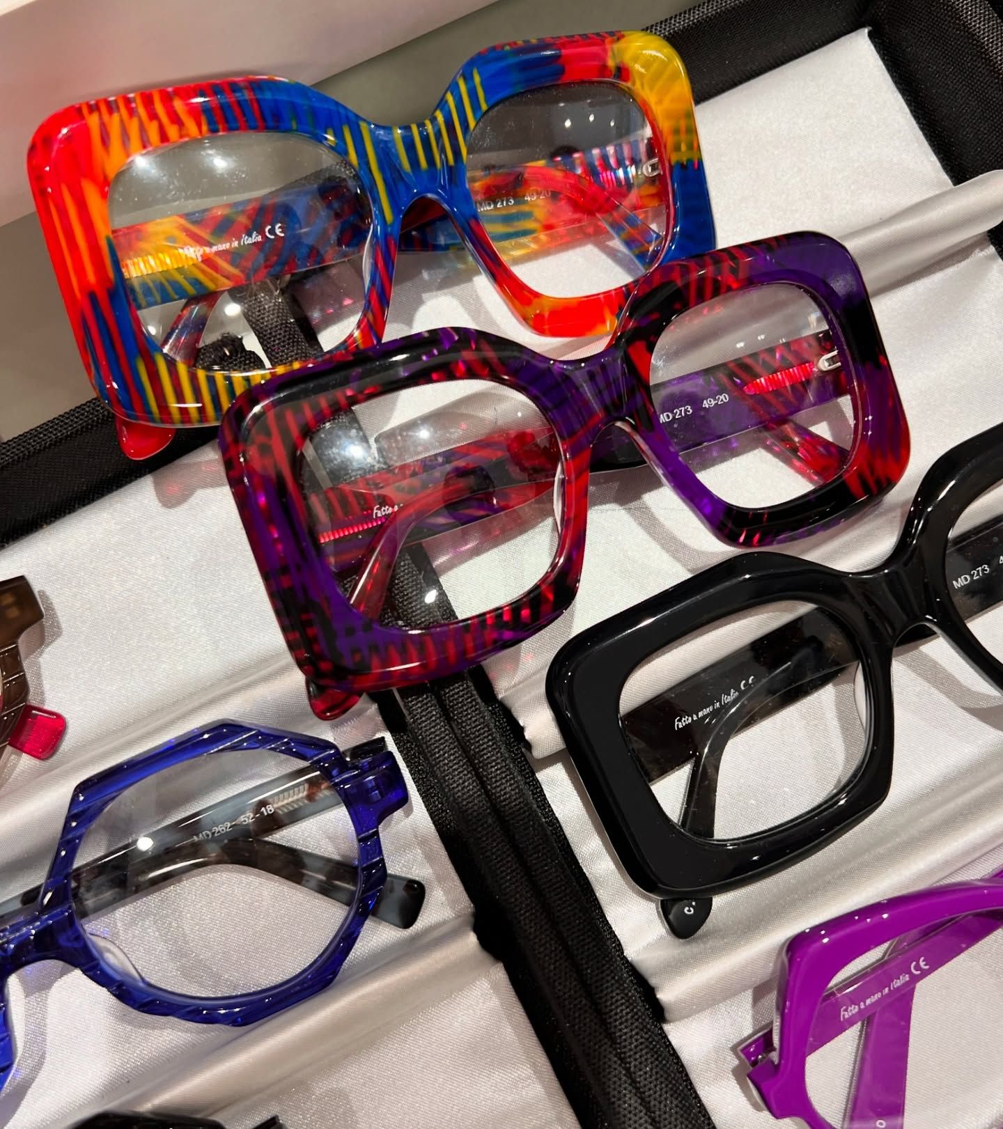 Una selezione di occhiali da vista colorati con montatura spessa, esposti in un astuccio foderato di velluto.