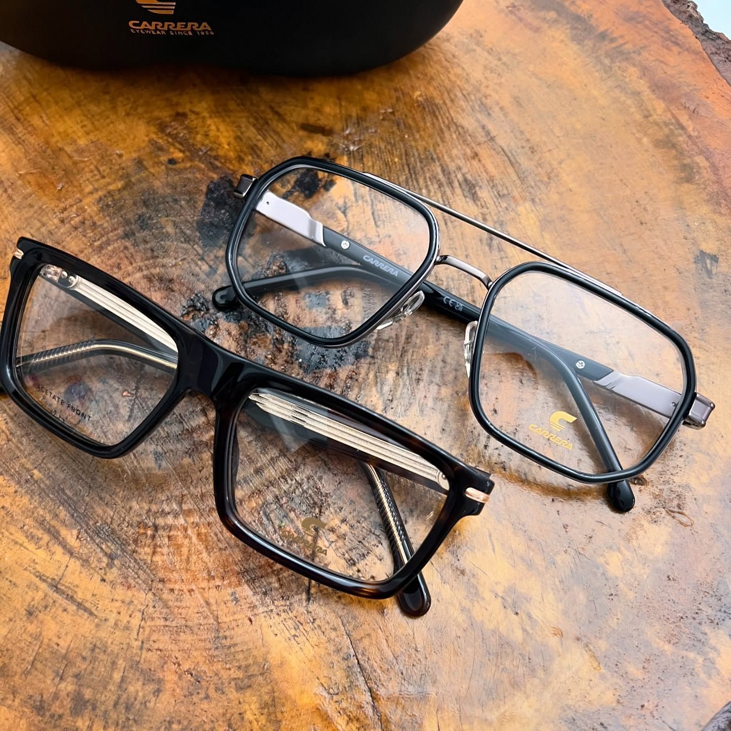 Due paia di occhiali da vista con montatura nera e aste metalliche poggiano su una superficie di legno ruvida accanto a un astuccio con il logo del marchio.