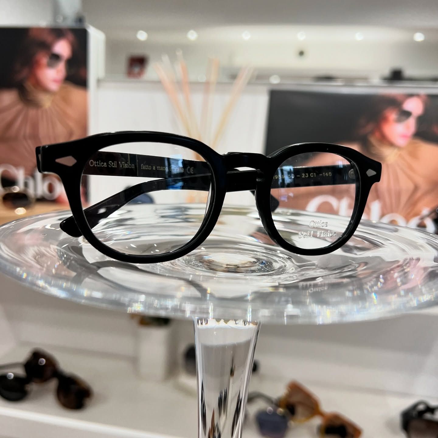 Occhiali da vista con montatura nera esposti su un supporto trasparente a piedistallo all'interno di un negozio.