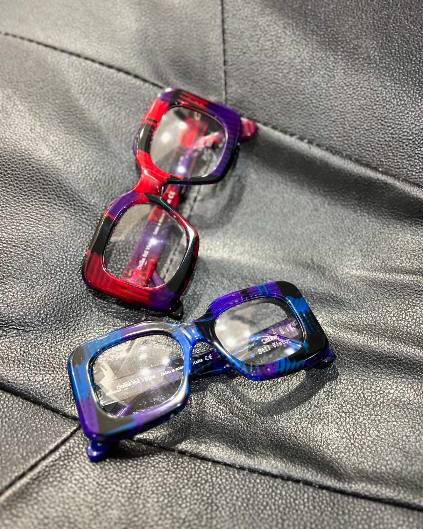 Due paia di occhiali dalla montatura spessa e rettangolare, con motivi vivaci e multicolori, poggiati su una superficie nera ruvida.