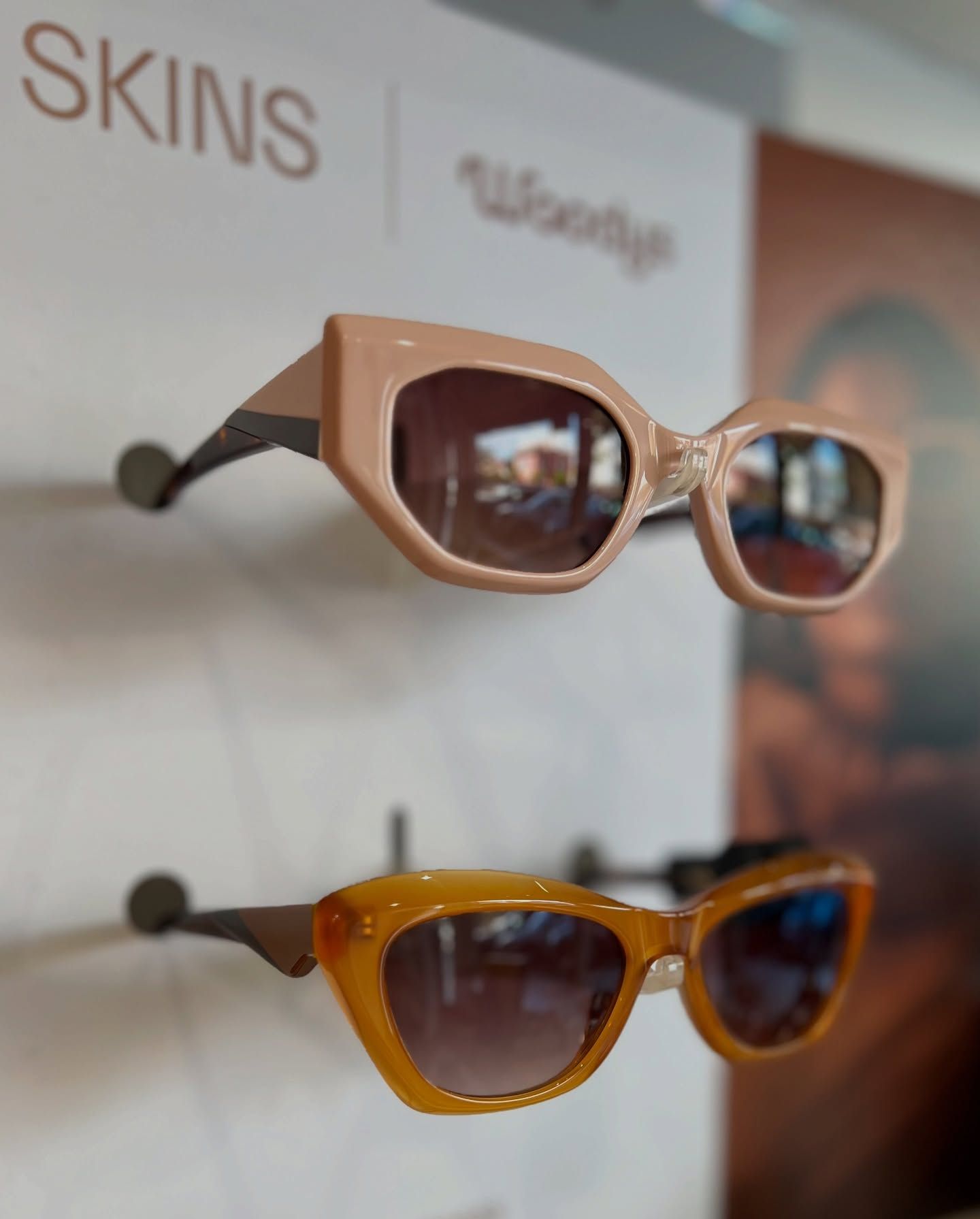 Due paia di occhiali da sole eleganti con montatura colorata, esposti su una parete bianca sotto l'etichetta del marchio 