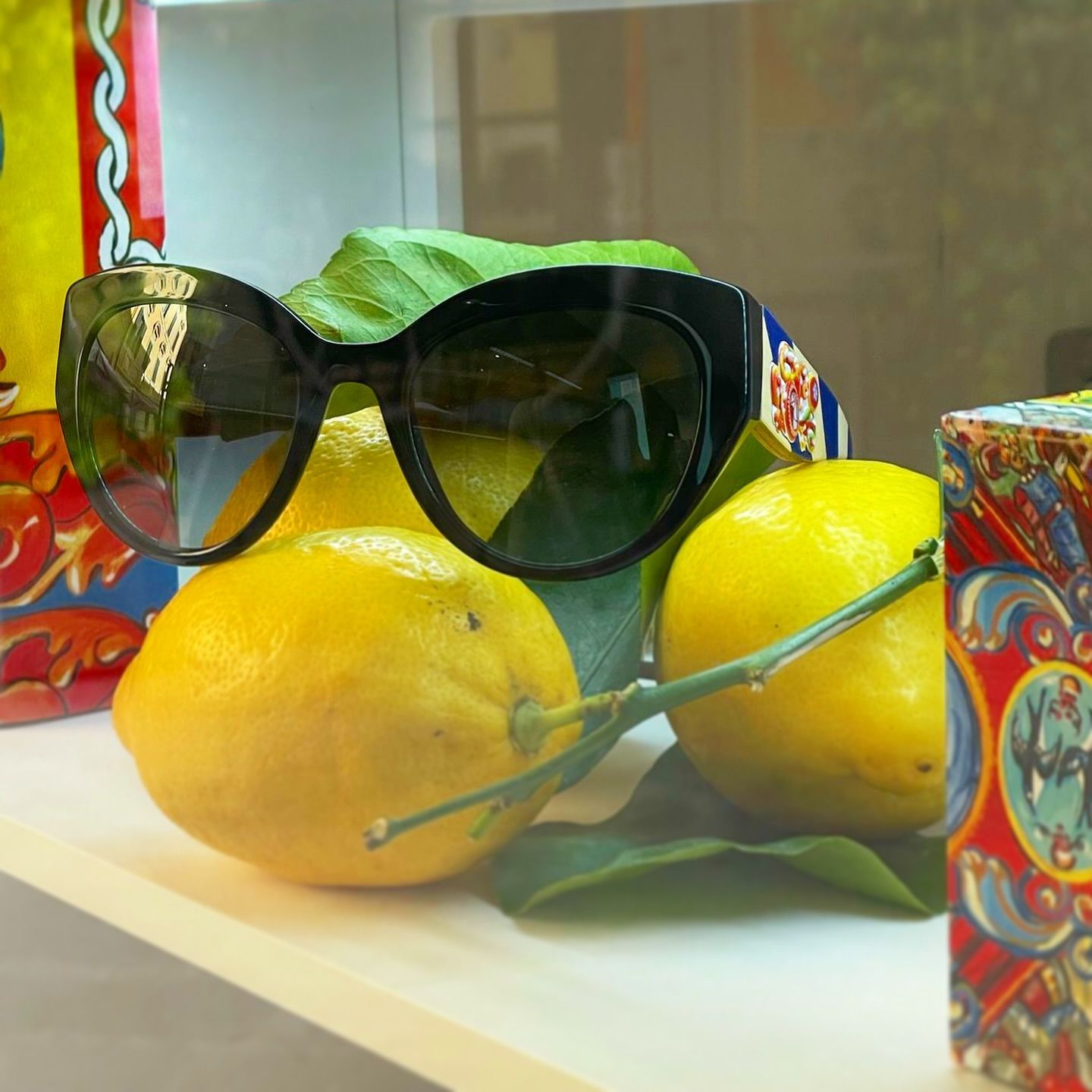 Occhiali da sole neri firmati, appoggiati su limoni giallo brillante con foglie verdi, esposti accanto a scatole colorate e decorate.
