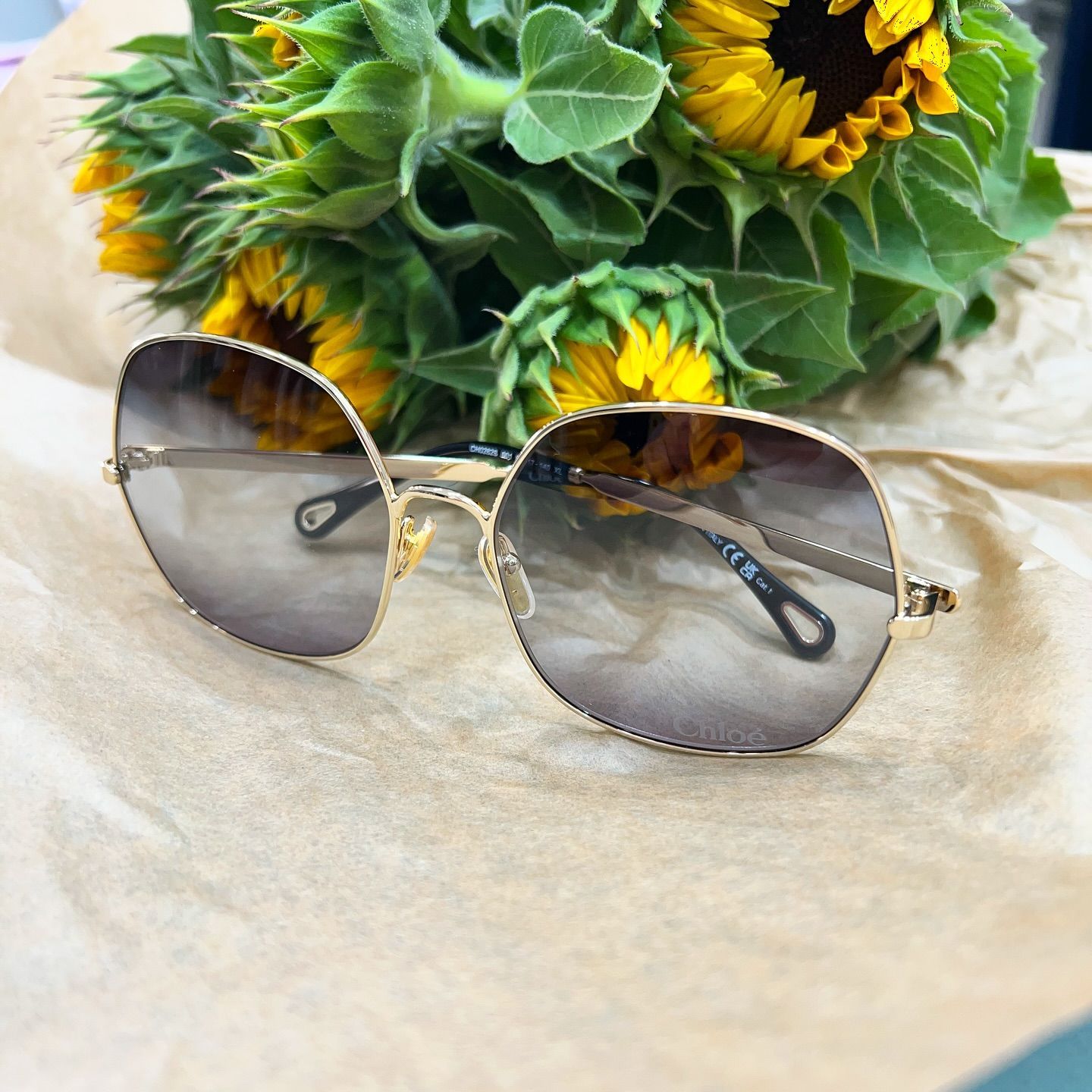 Occhiali da sole con montatura dorata e lenti sfumate appoggiati su un foglio di carta marrone davanti a un mazzo di girasoli.