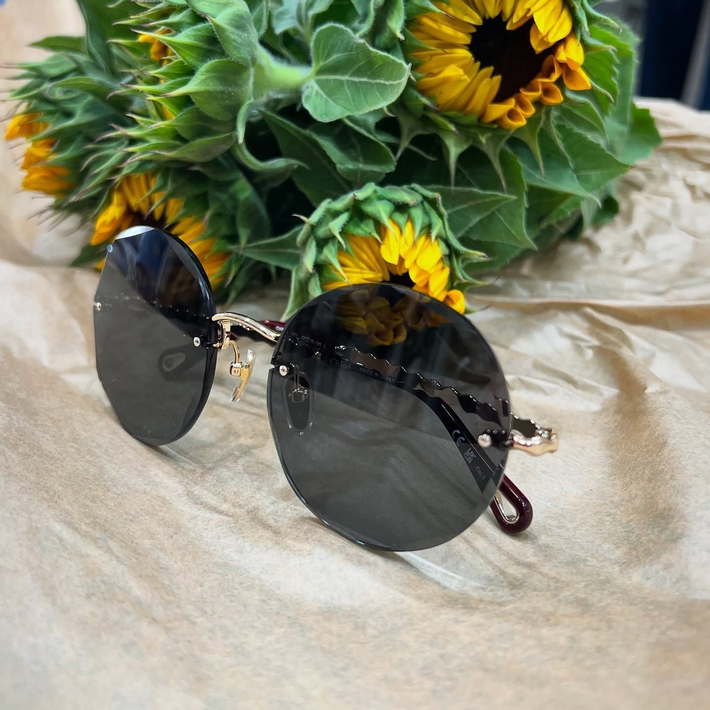 Occhiali da sole scuri e rotondi con montatura sottile dorata, appoggiati su carta marrone ruvida davanti a un mazzo di girasoli.