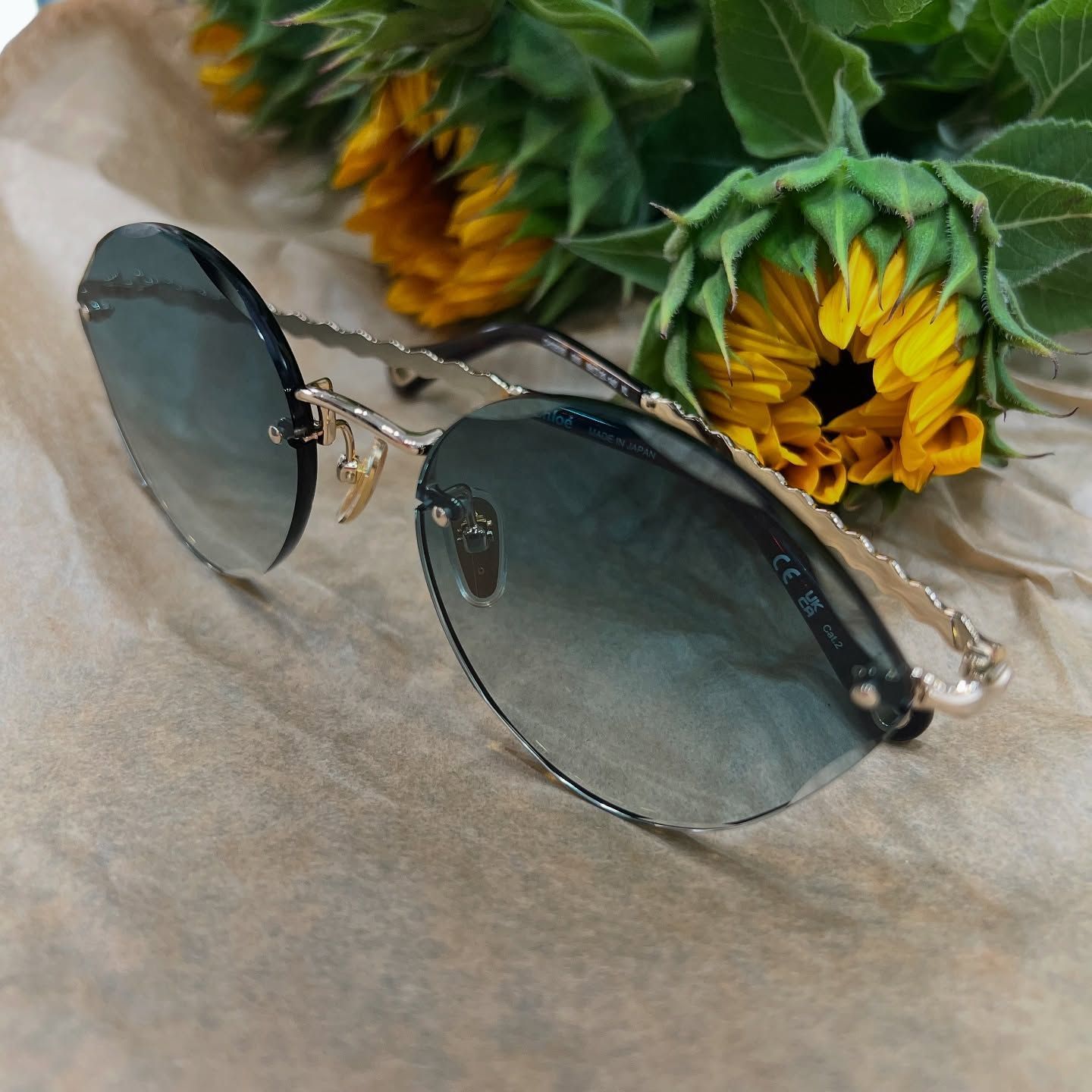 Occhiali da sole senza montatura con aste dorate e testurizzate e lenti blu sfumate poggiano su un foglio di carta beige stropicciato accanto a dei girasoli.