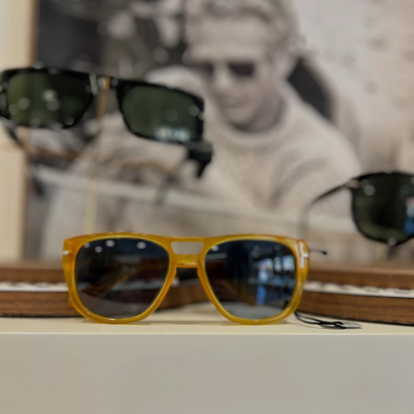 In primo piano si vedono occhiali da sole color ambra con montatura nera sfocata e, sullo sfondo, il ritratto di una persona.