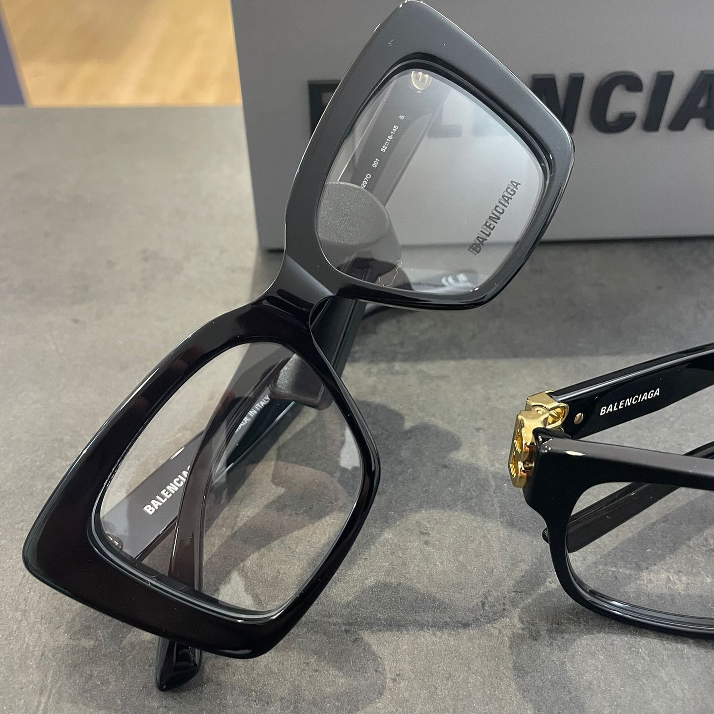 Due paia di occhiali da vista Balenciaga neri su una superficie grigia, con una scatola con il logo del marchio sullo sfondo.
