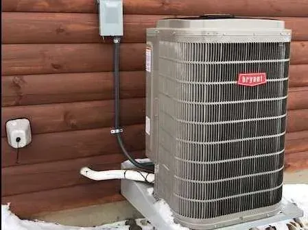 Bryant air conditioning unit on a snow-covered platform next to a brown wooden building. Electrical components are visible.