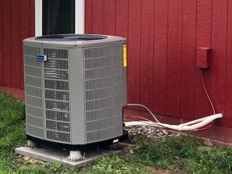 An air conditioning unit against a red building, on a concrete pad, with white tubing.