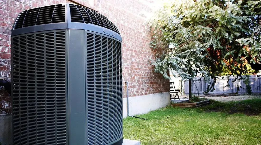 Gray air conditioning unit next to a brick wall and a grassy yard with a tree.