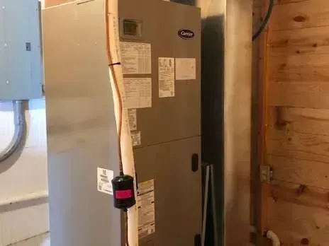 A gray Carrier HVAC unit in a room with wood paneling and a metal duct. A pink and black float switch is visible.