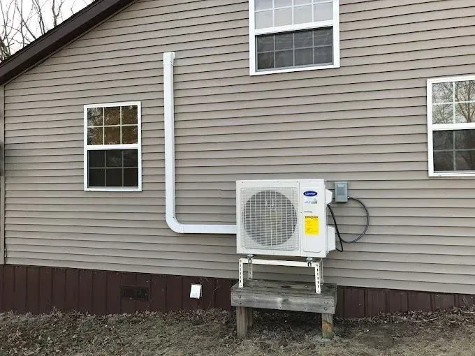Air conditioning unit mounted on a wooden platform outside a house with beige siding and white windows.