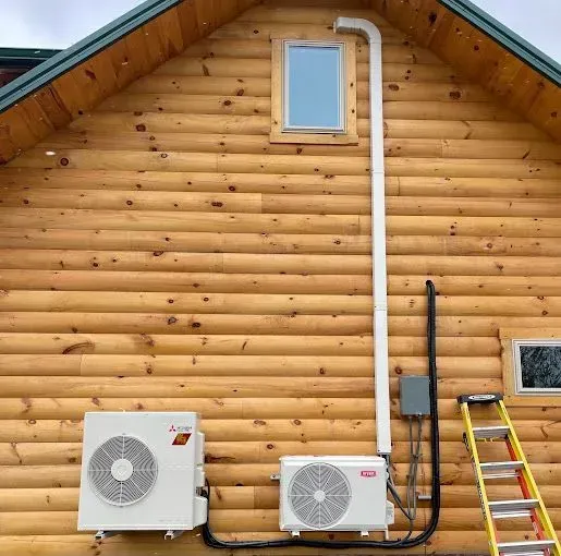 Two white AC units on a wood cabin wall with conduit. A ladder leans against the wall.