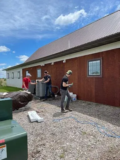 Three people installing an AC unit outside a building with brown siding and a metal roof on a sunny day.