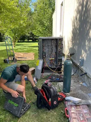 HVAC technician working on an air conditioning unit outside a house on a sunny day. Tools and equipment are nearby.