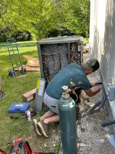 HVAC technician repairs an air conditioning unit outside a building, using tools and a gas cylinder.