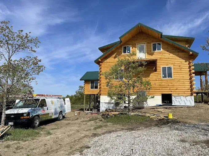 Log cabin under construction with a white van parked on gravel, under a blue sky.