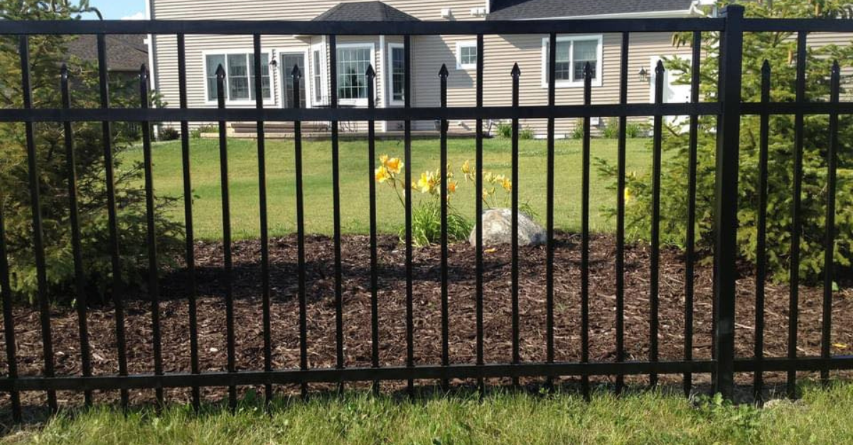 Black metal fence in front of a house, with green grass and trees in the background.