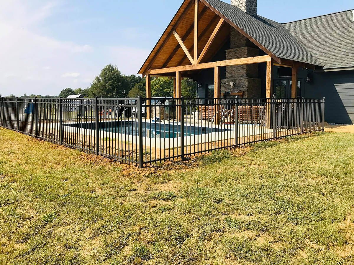 A backyard swimming pool enclosed by a black metal fence next to a wooden outdoor patio structure with a stone fireplace.
