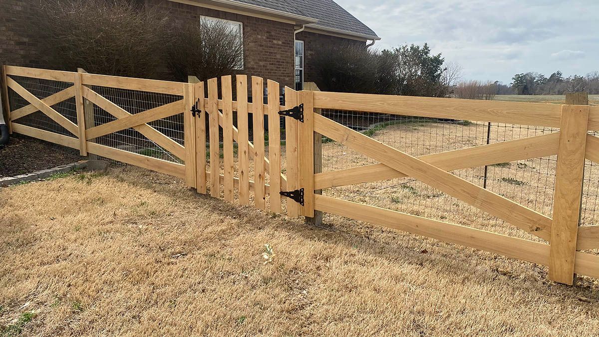 A wooden split-rail fence with an arched gate in front of a brick house on a grassy lawn.