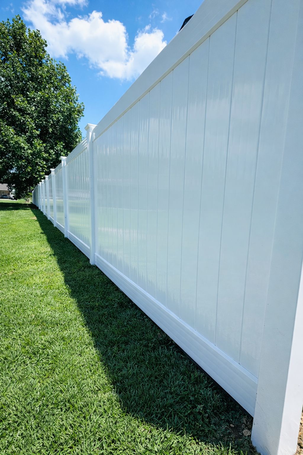 A tall, white vinyl privacy fence lines a lush green lawn under a bright blue sky with light clouds.