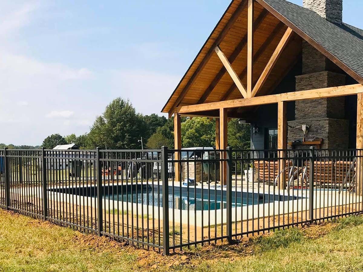 A black metal fence surrounds a swimming pool next to a wooden outdoor pavilion with a stone fireplace.
