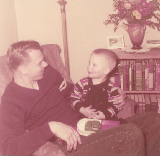 Man and child in a chair. Man in dark shirt, child in striped shirt, toy visible. Bookshelf, vase of flowers in background.
