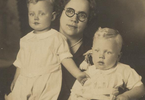 Woman with round glasses and pearl necklace holds two young children in white outfits.