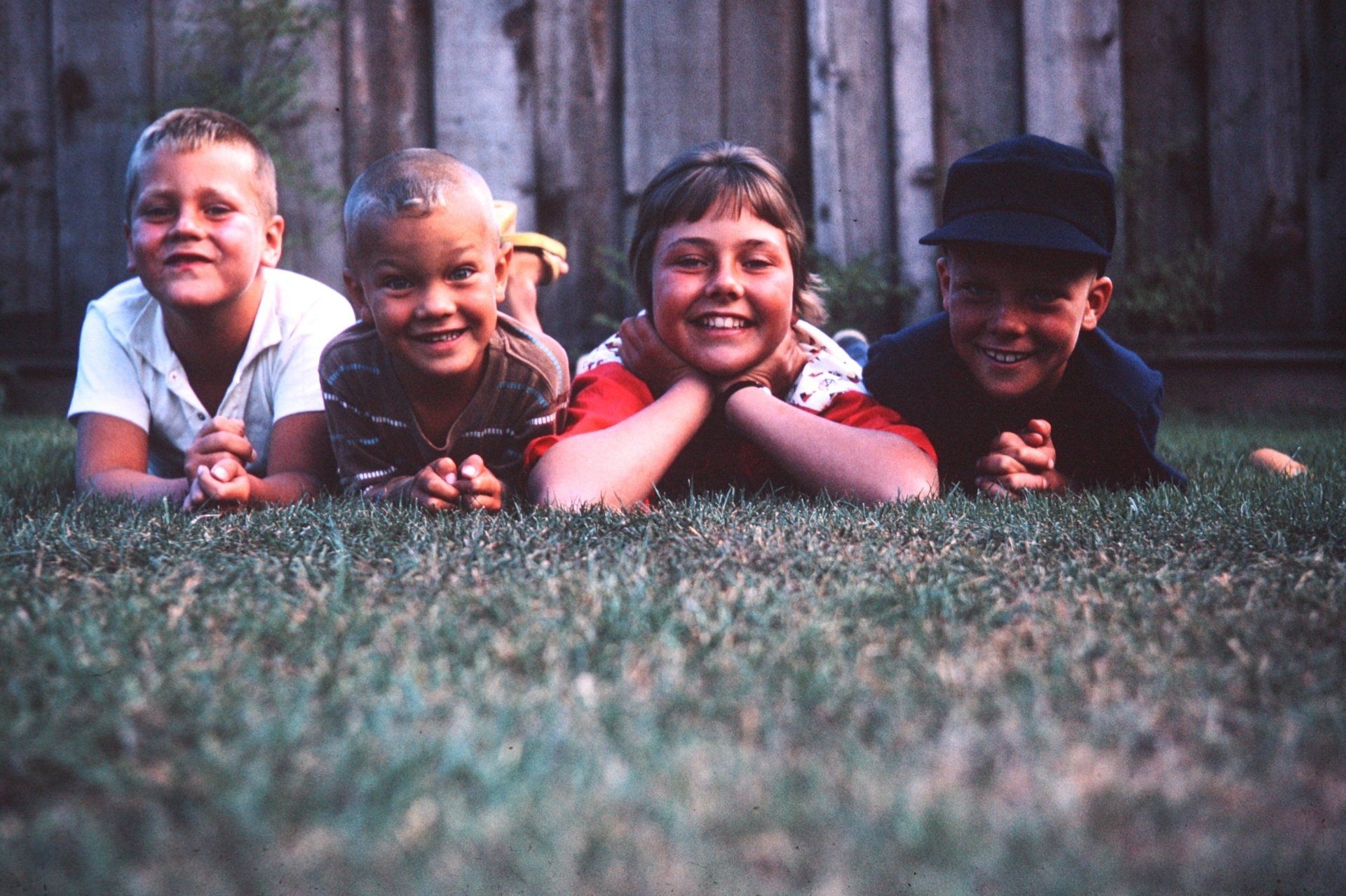 Four smiling children lying on grass in front of a wooden fence.