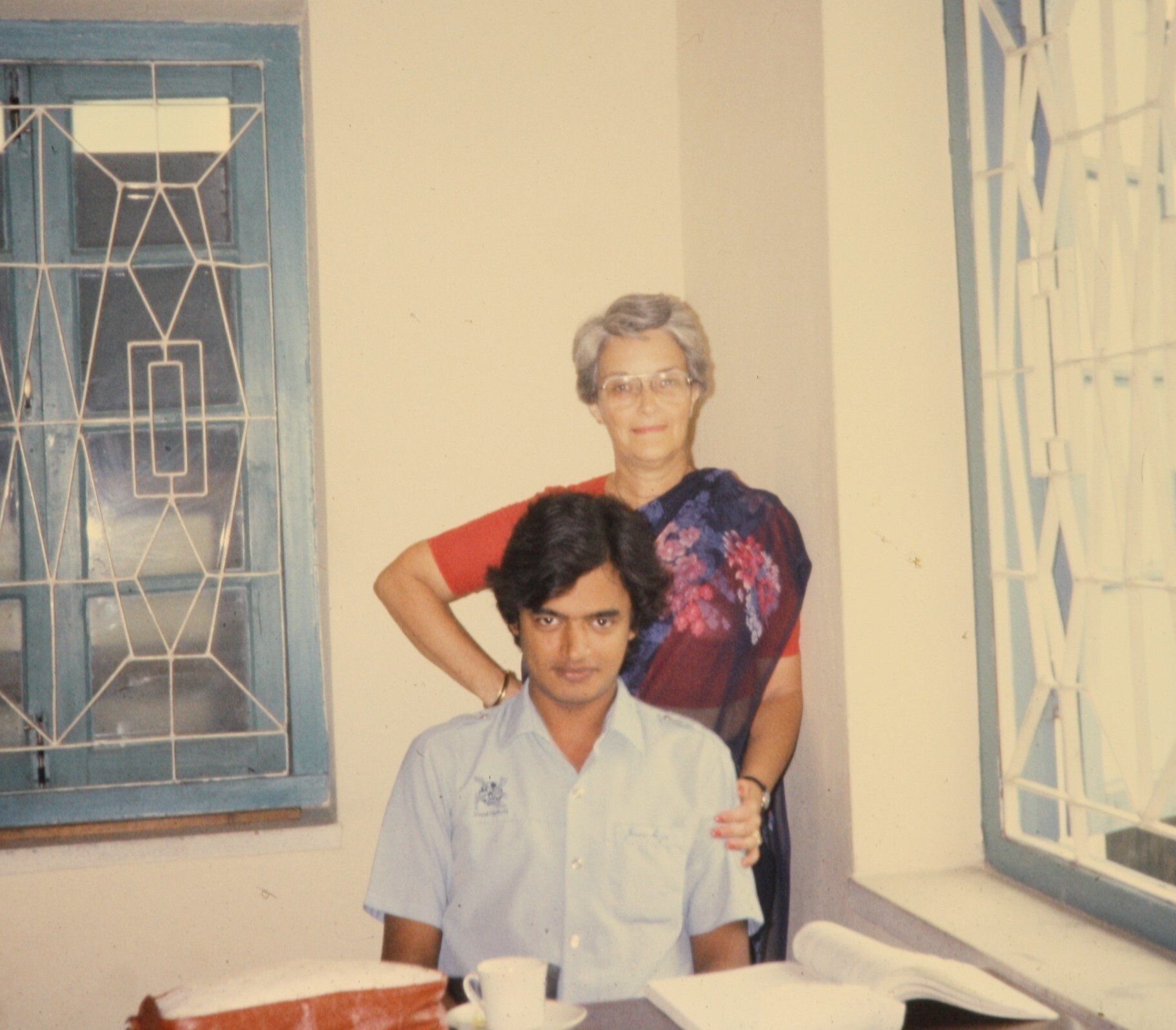 Man sitting at a desk, woman standing behind him with arm on his shoulder, by a window.