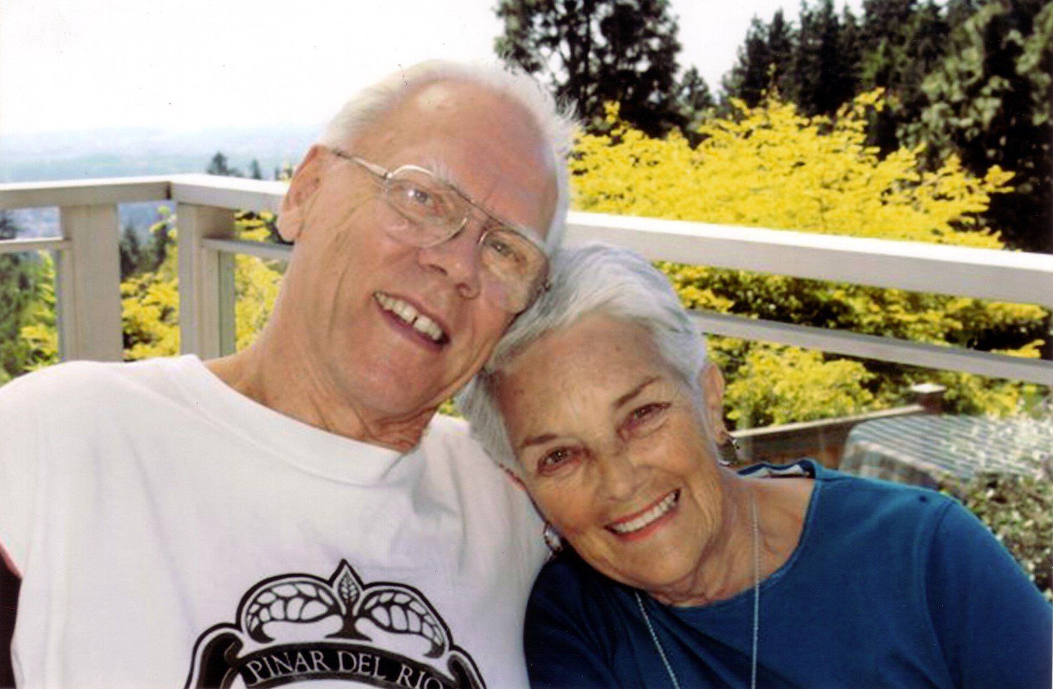 Smiling elderly couple outdoors, woman resting head on man's shoulder; trees and railing in the background.