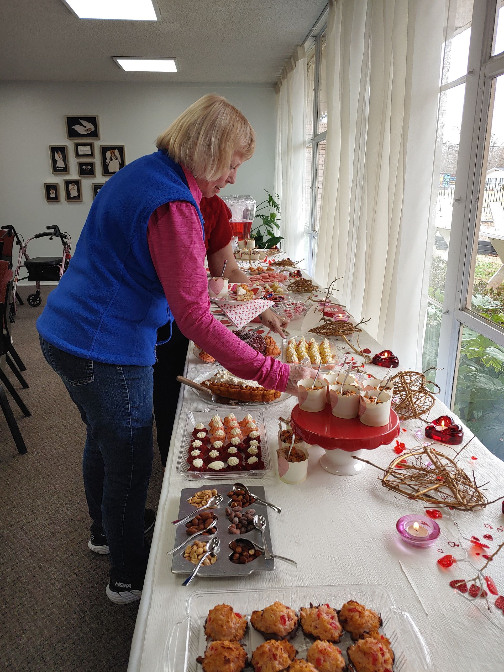 Woman in blue vest at a table laden with sweets near a window.