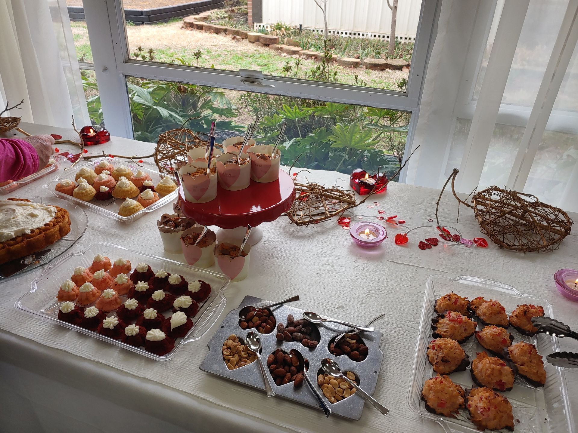 Dessert table with various sweets, red and white color scheme. Cakes, cupcakes, and cookies are displayed with garden view.