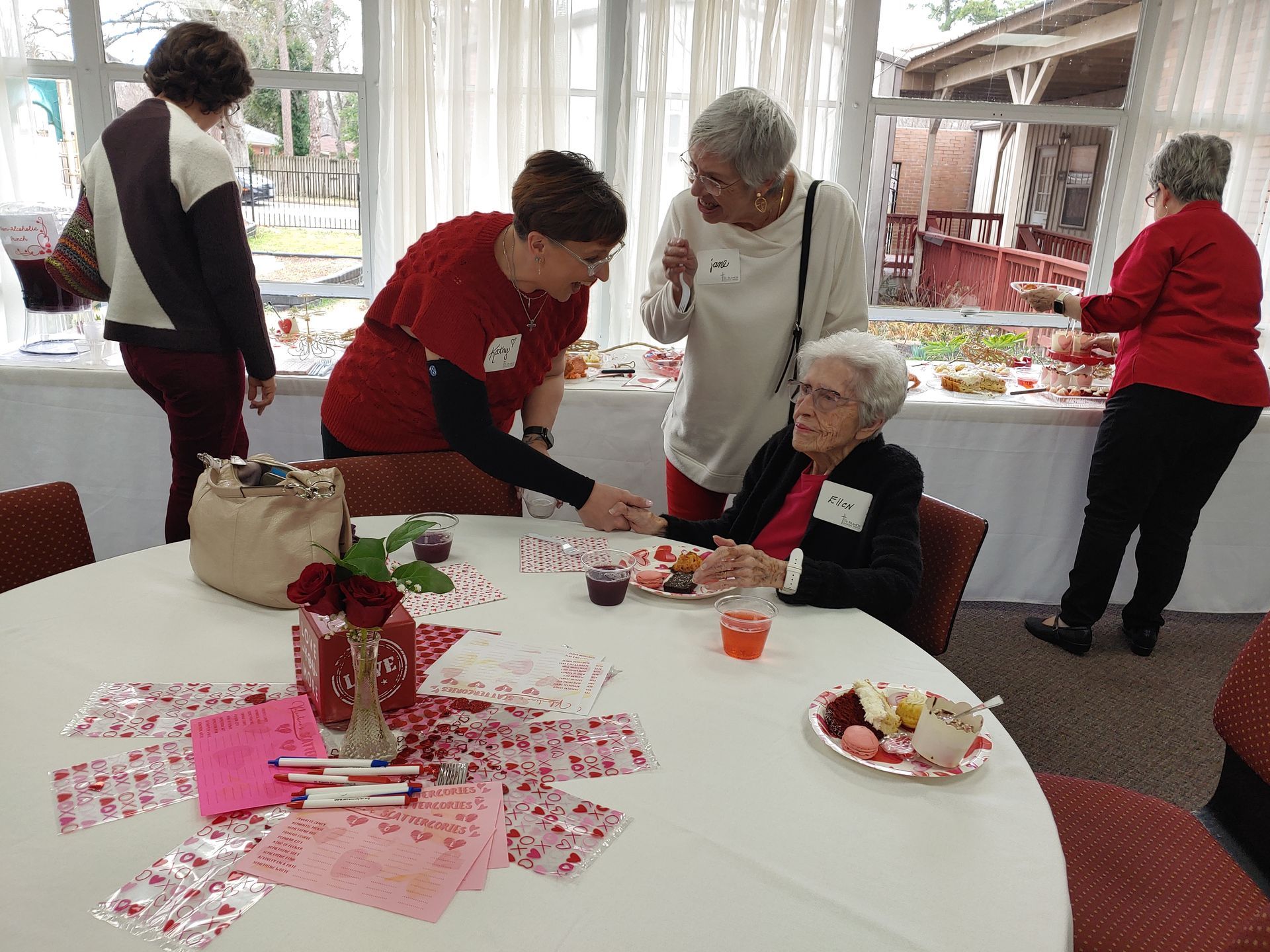 People gathered around a table with Valentine's Day decorations and treats. Some look at food, others talk.