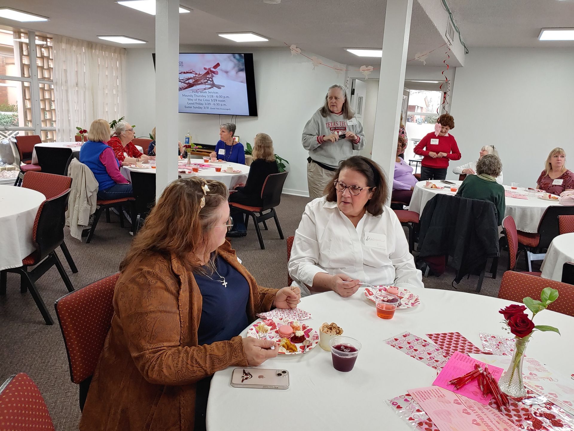 People at round tables in a decorated room, some eating, others talking. A woman gives a presentation.