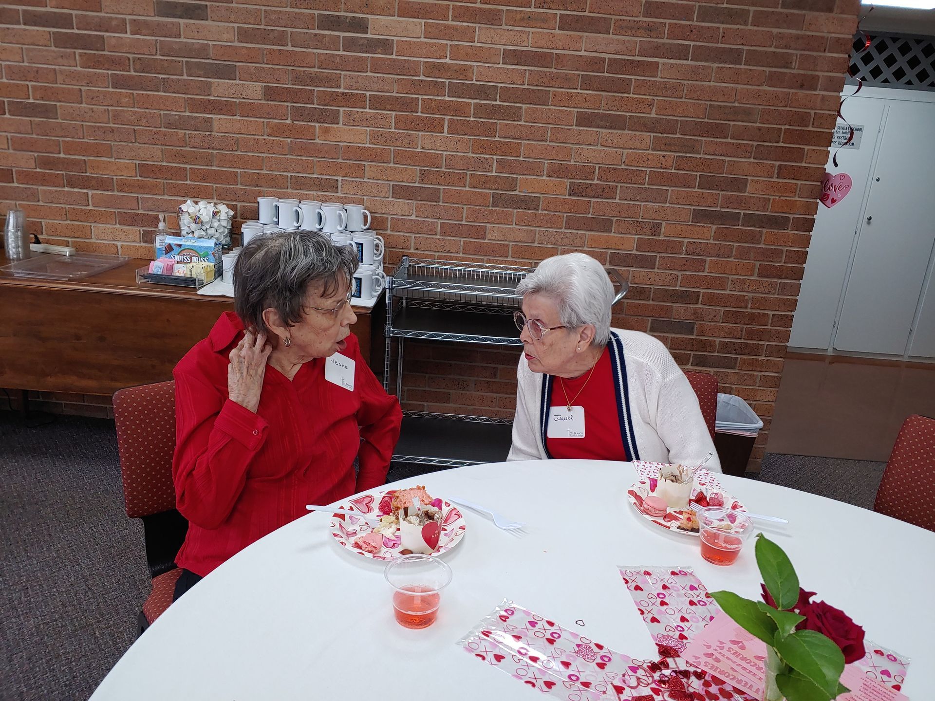 Two women sit at a decorated table, chatting. Red and white accents, dessert plates. Brick wall background.
