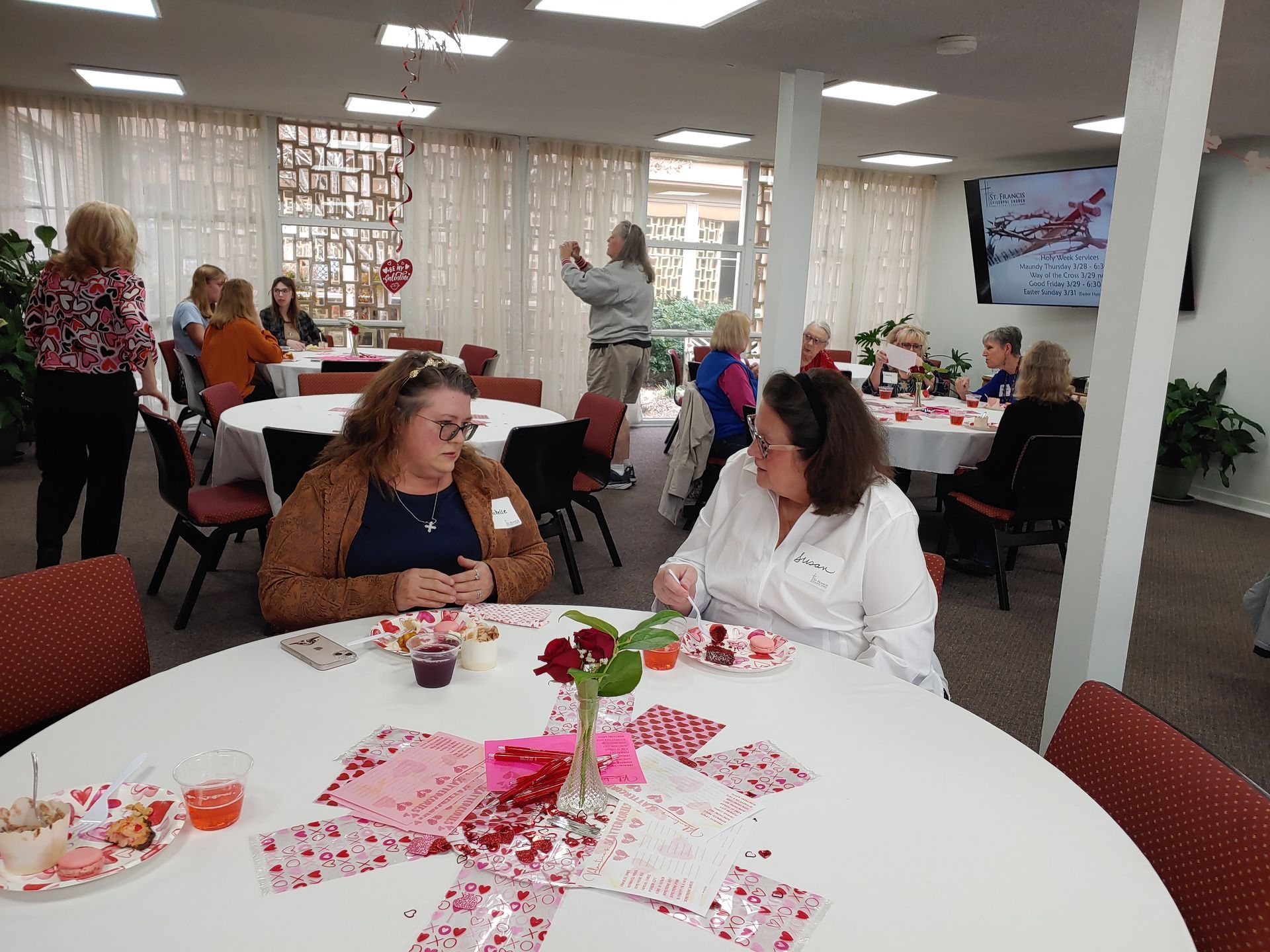 People at round tables in a decorated room, some talking, holding beverages. Valentine's Day decorations.
