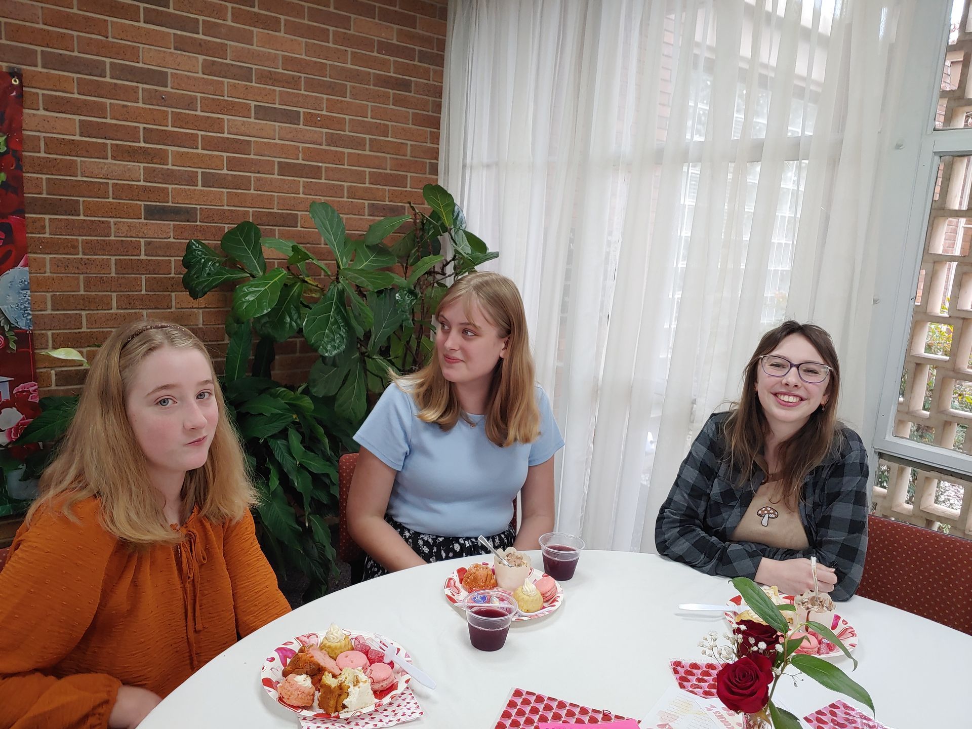 Three women seated at a round table with food and drinks; brick wall and large plant in background.
