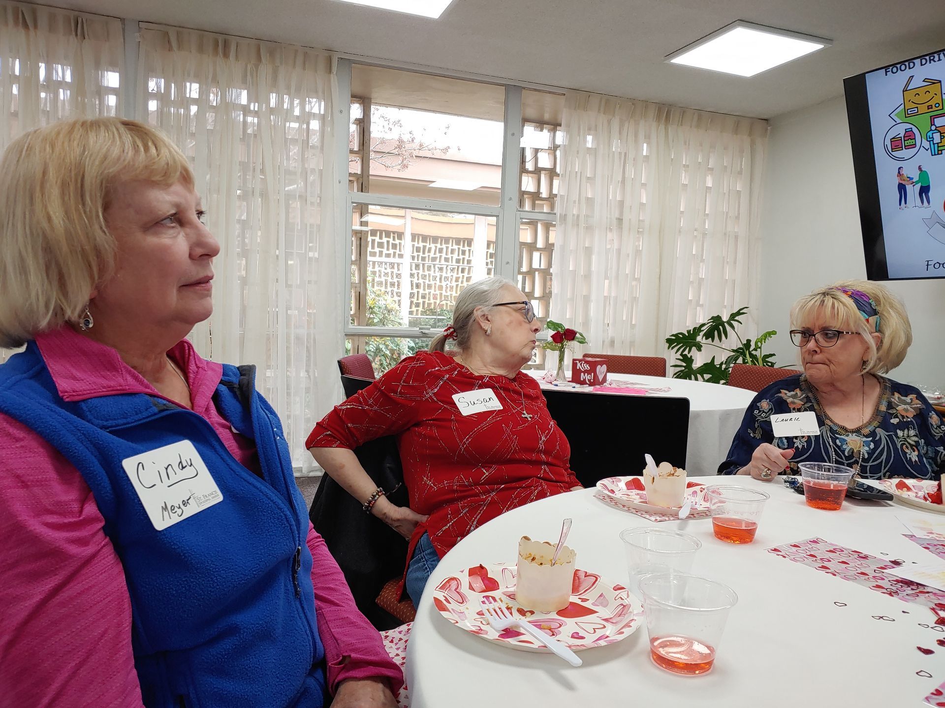 Three women seated at a table, attending an event. One wears a blue vest, another a red shirt, and the third a patterned blouse.