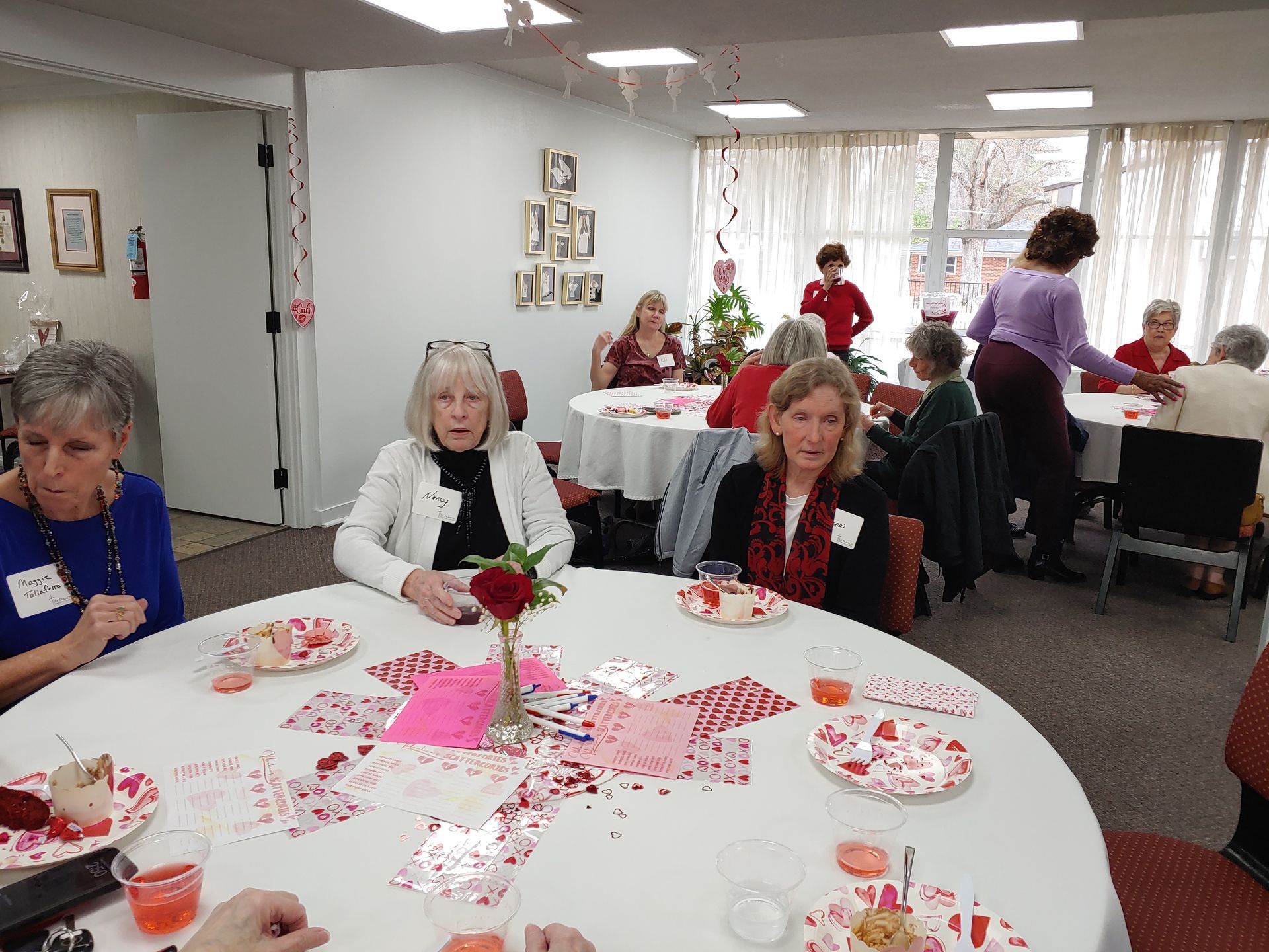 Women at tables decorated for Valentine's Day. Some seated, others interacting near a window. Red accents throughout the room.