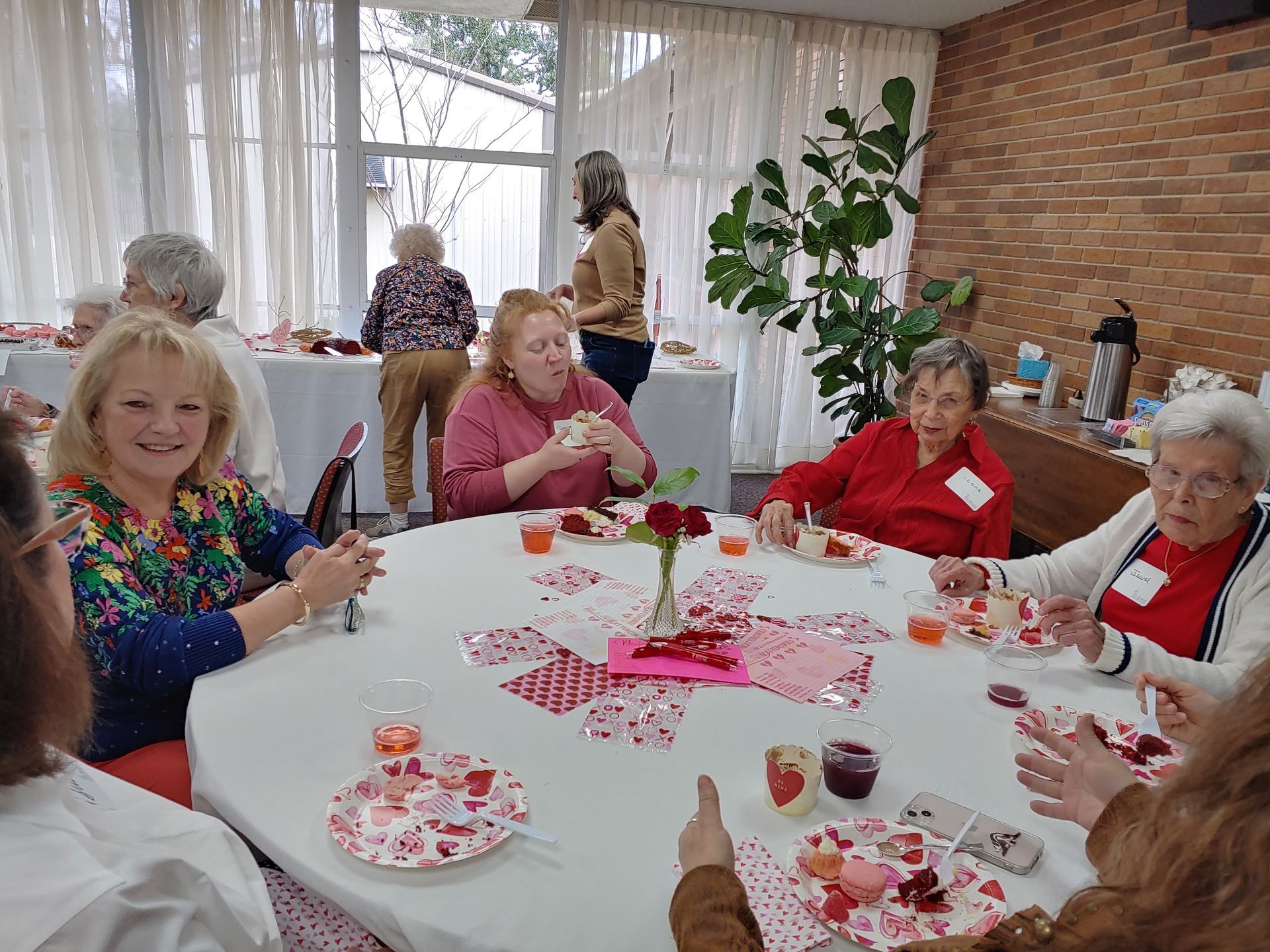 People at a Valentine's Day party seated around a decorated table, eating and drinking. Brick wall and plant in background.