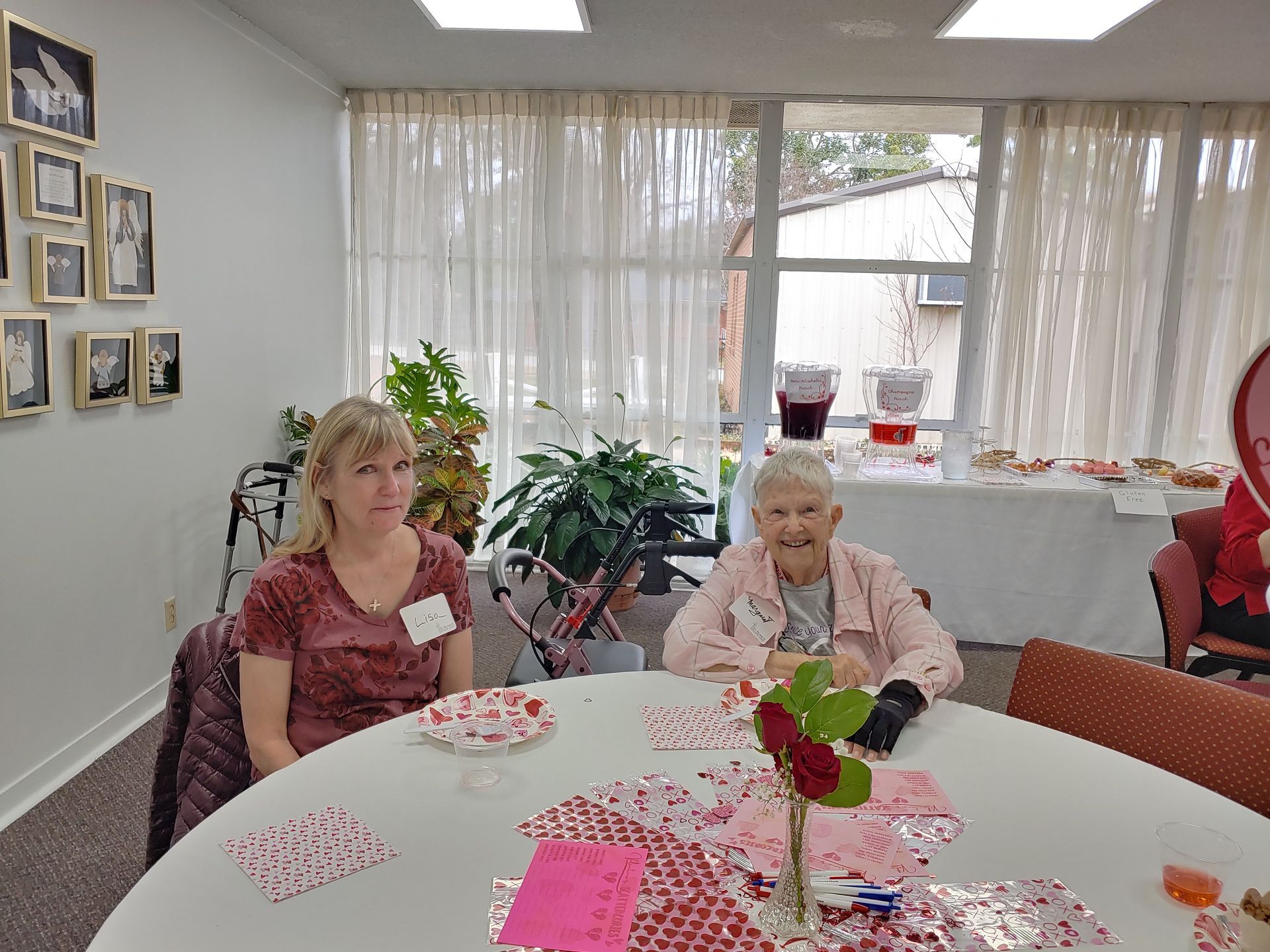 Two women sit at a decorated table, possibly at a social gathering. A plant and refreshments are nearby.