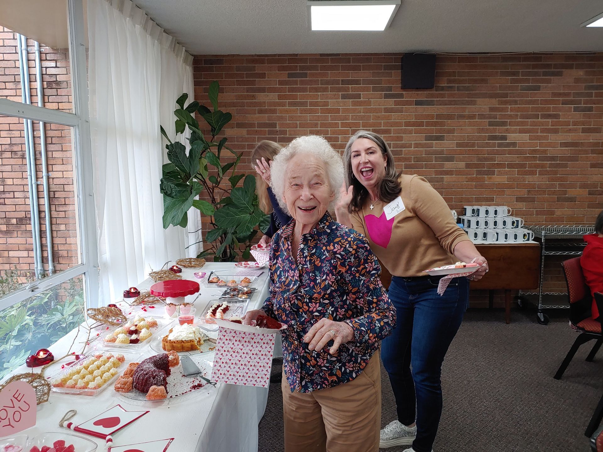 Two smiling women at a Valentine's Day dessert table, one older, with treats, in a room with a brick wall.