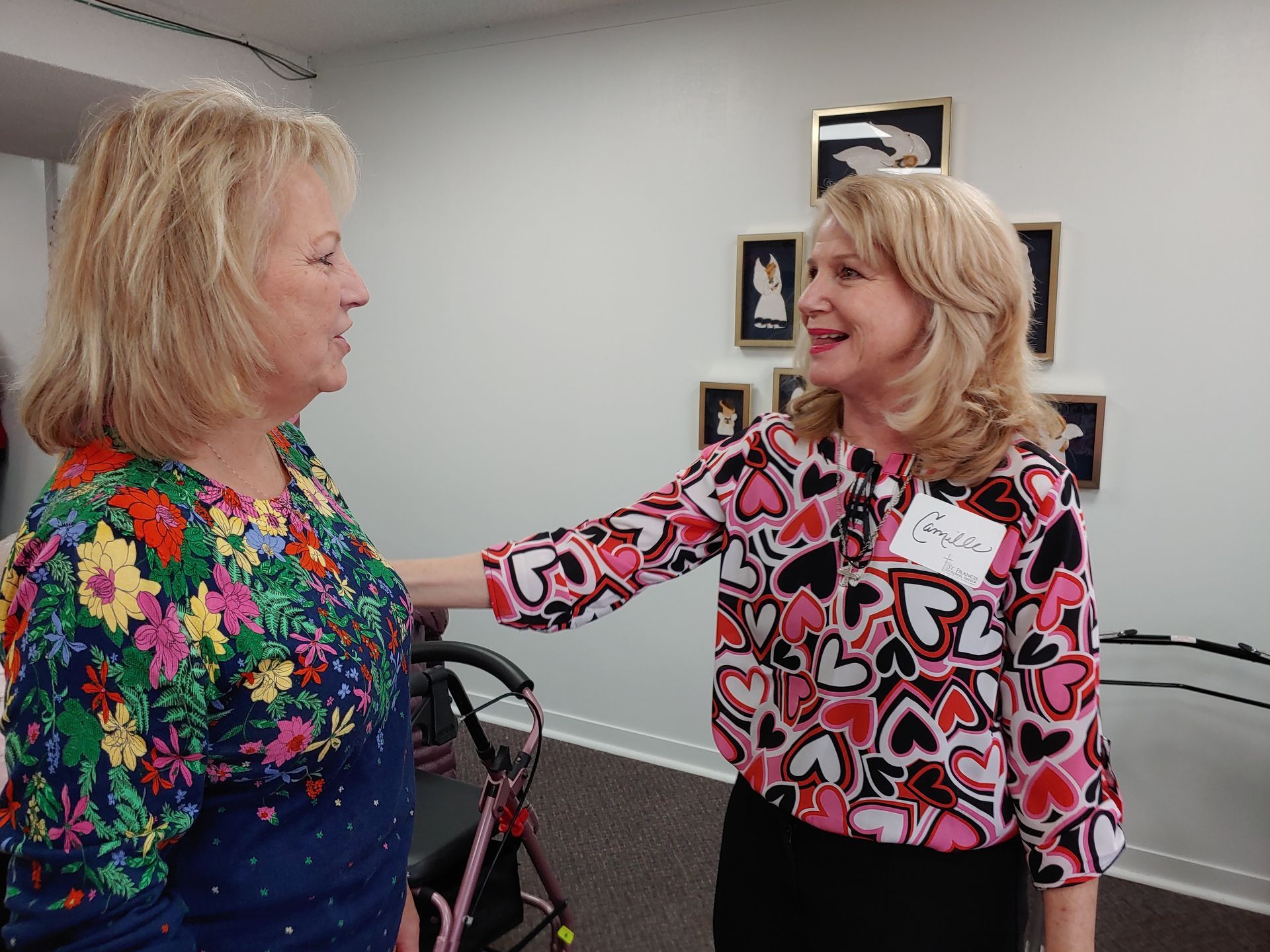 Two women in a room, one with hand on the other's shoulder. Wall art behind them.
